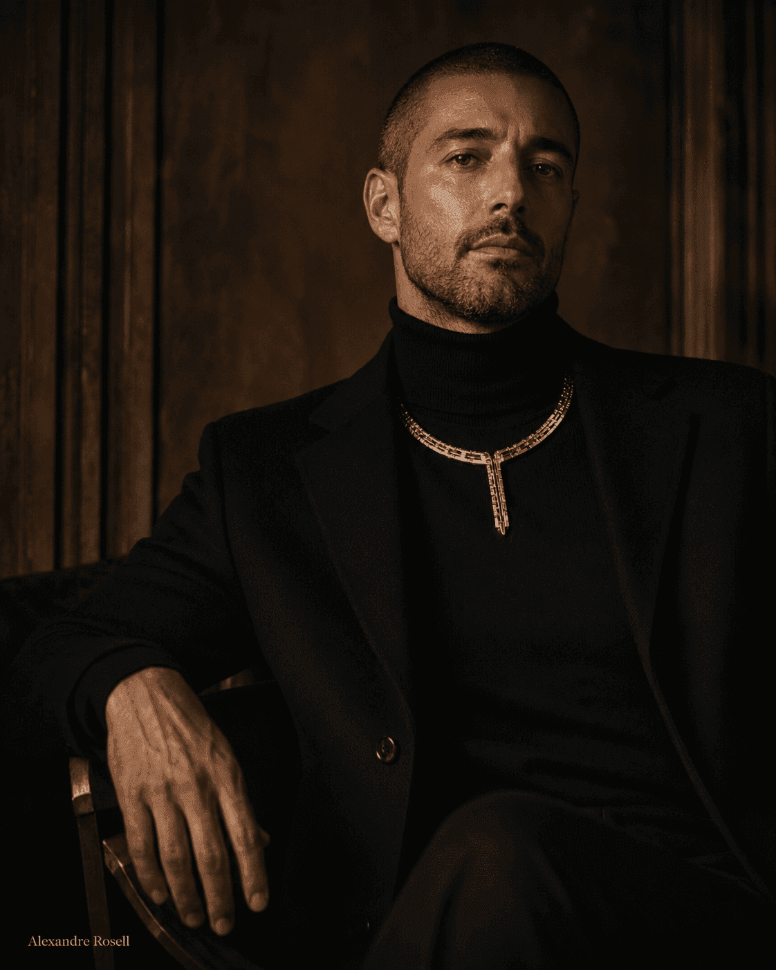 A man in black wearing a gold chain, seated against panelled wood.