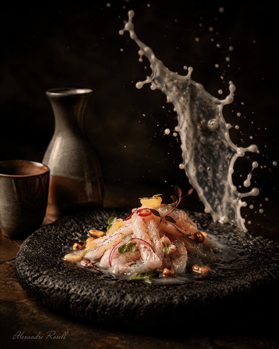 Cured fish dressed on dark volcanic stone, a ribbon of liquid suspended mid-splash beside a sake vessel.