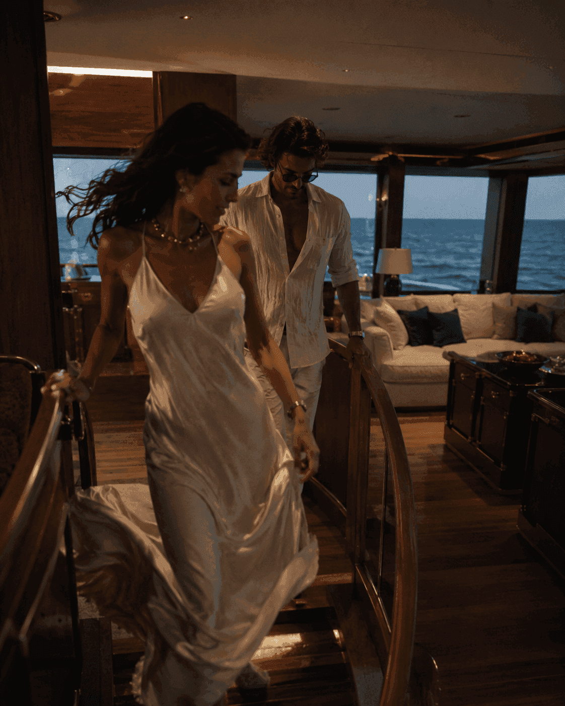 A couple in white descending into the warm-lit saloon of a yacht, the open sea through the windows.