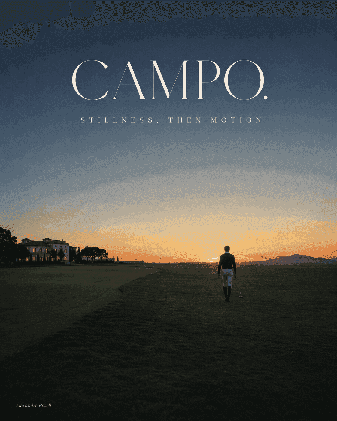 A lone figure crossing the field toward a lit clubhouse as the sun sets behind the hills.